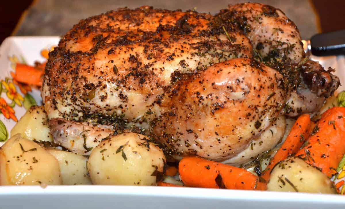 Golden roasted chicken covered with herbs on a bed or roasted potatoes and carrots.