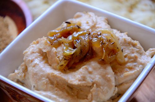 Hummus in a square pot with caramelized onion garnish.
