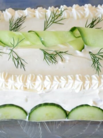 Rectangluar sandwich loaf, frosted with cream cheese and mayonnaise, piped around the edges and garnished with cucumber slices and fresh dill fronds.