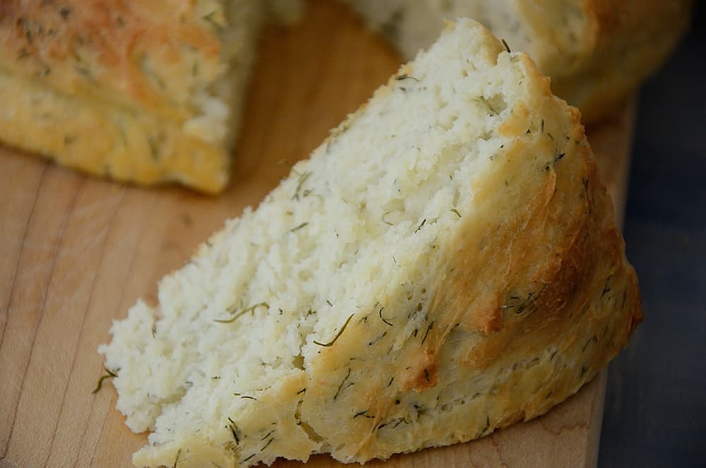 A wedge of no knead dill bread on a cutting board with the loaf behind it.