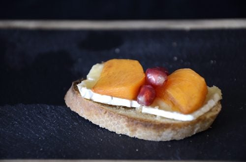 Persimmon slices and pomegranate seeds on melted Brie on baguette slice.
