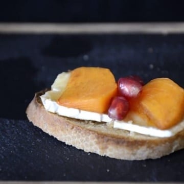 Persimmon slices and pomegranate seeds on melted Brie on baguette slice.