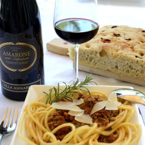 A pasta bowl with Bigoli covered with duck ragu, garnished with grana padano and rosemary at a table with a glass of amarone wine and focaccia bread.