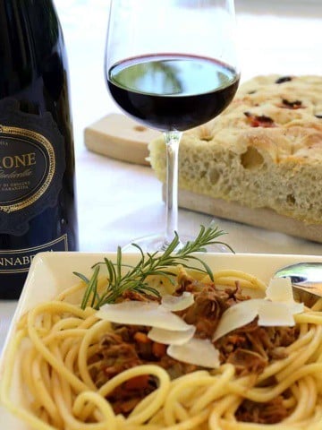 A pasta bowl with Bigoli covered with duck ragu, garnished with grana padano and rosemary at a table with a glass of amarone wine and focaccia bread.