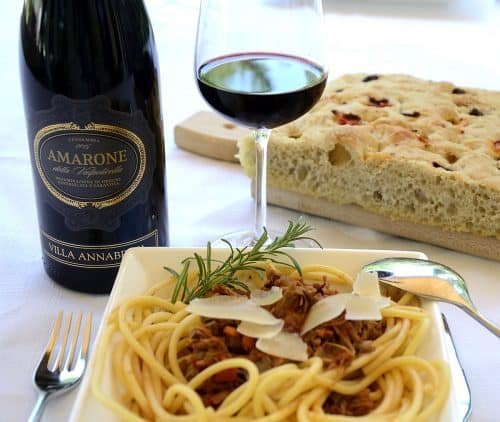 A pasta bowl with Bigoli covered with duck ragu, garnished with grana padano and rosemary at a table with a glass of amarone wine and focaccia bread.