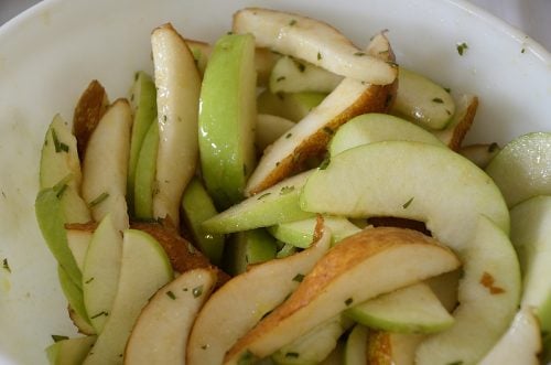 Slices of apples and pears in a bowl tossed in olive oil and minced rosemary.