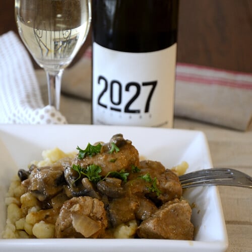 Place setting with a dish of juicy, creamy cubes of cooked pork and mushrooms over German Spatzle noodles and a glass of white wine on the side.