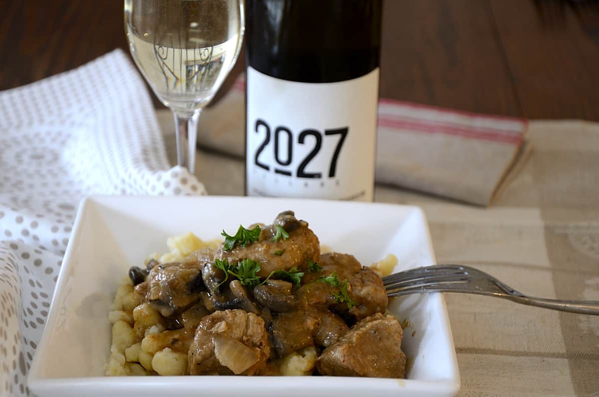 Place setting with a dish of juicy, creamy cubes of cooked pork and mushrooms over German Spatzle noodles and a glass of white wine on the side.