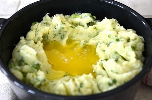 Casserole of mashed potatoes with green onions and parsley mixed in and a well of melted butter in the center.