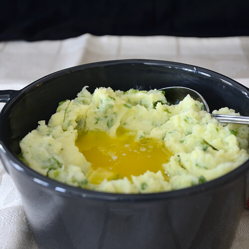 Casserole of mashed potatoes with green onions and parsley mixed in and a well of melted butter in the center.