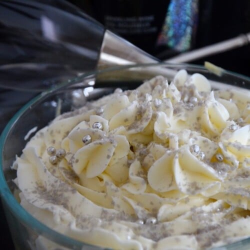 Bowl of champagne trifle with piped icing and silver balls and fleck to decorate. Champagne bottle and flutes are in the background.