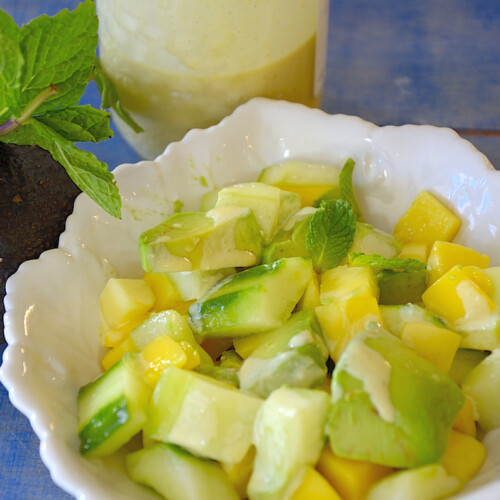 Chopped avocado, mango and cucumber in a salad bowl with creamy Dijon Vinaigrette drizzled on top.