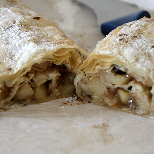 Flaky apple strudel with phyllo pastry topped with icing sugar cut open.