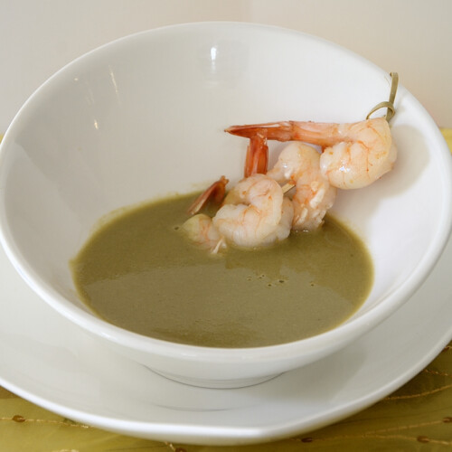 Asparagus soup in a white bowl with a skewer of shrimp.