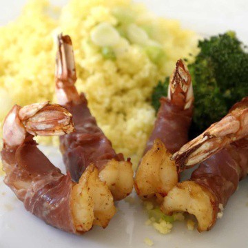Jumbo shrimp wrapped in prosciutto on a plate with broccoli and couscous.