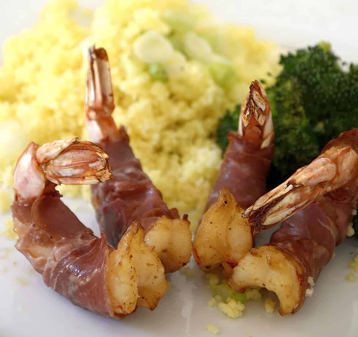 Jumbo shrimp wrapped in prosciutto on a plate with broccoli and couscous.