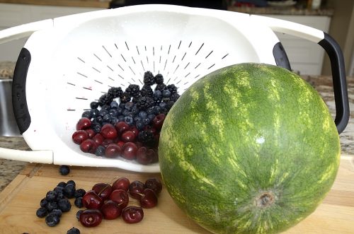 A whole watermelon on a cutting board with fresh berries spilling out of a colander beside.
