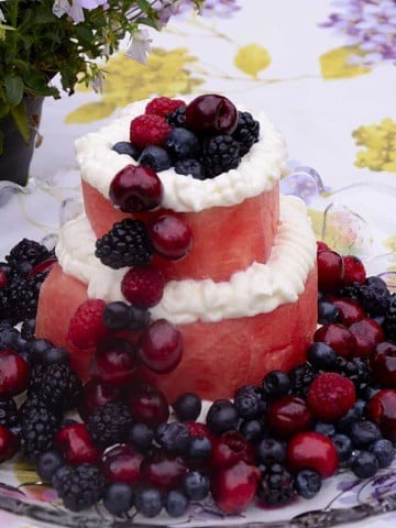 Two tiers of watermelon with cream cheese frosting piped around the edge of each layer and cherries, blackberries, blueberries and raspberries pile on the top, down one side and round the base.