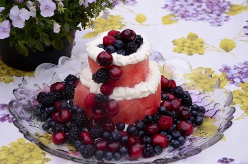 Two tiers of watermelon with cream cheese frosting piped around the edge of each layer and cherries, blackberries, blueberries and raspberries pile on the top, down one side and round the base.