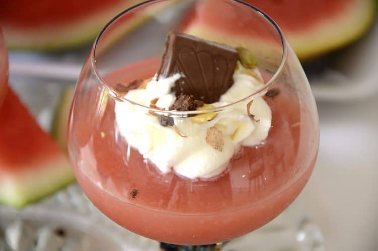 Stemmed cup with watermelon pudding topped with whipped cream and garnished with crushed pistachios and chocolate shavings.