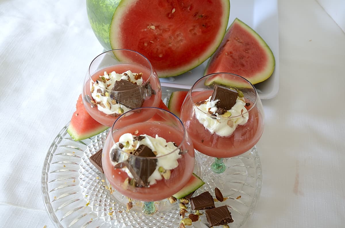 Stemmed cups with watermelon pudding topped with whipped cream and garnished with crushed pistachios and chocolate shavings