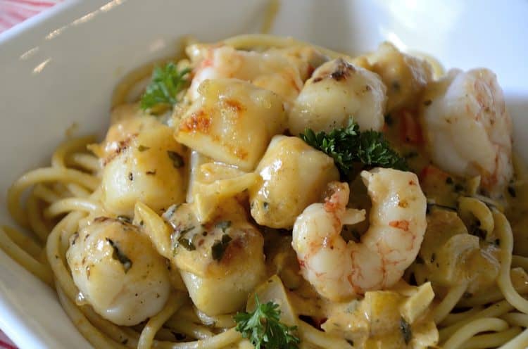 Close up of jumbo shrimp and scallops in a creamy sauce with parsley and sun-dried tomatoes over a bowl of spaghetti.