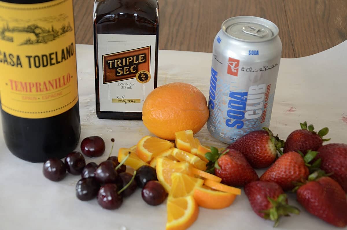 Bottle of tempranillo wine, Triple Sec, Club Soda, oranges, oranges slices, strawberries and cherries on a board.