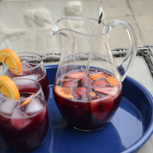 Pitcher of Sangria with red wine, oranges slices, strawberry slices and cherry slices. Two glasses on the side filled and garnished with orange slices.