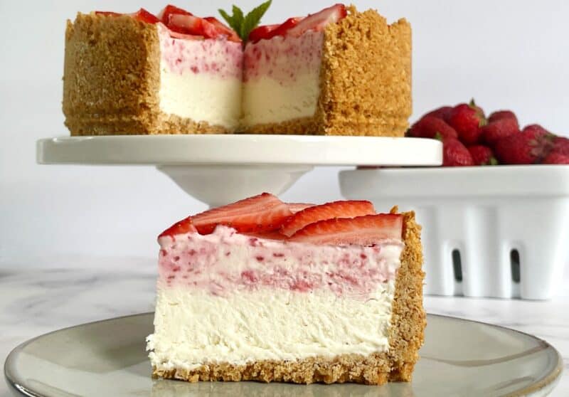 Strawberry ice cream cake cut with a piece on a plate showing 2 layers of ice cream, one vanillla, one strawberry in a graham cracker crust.