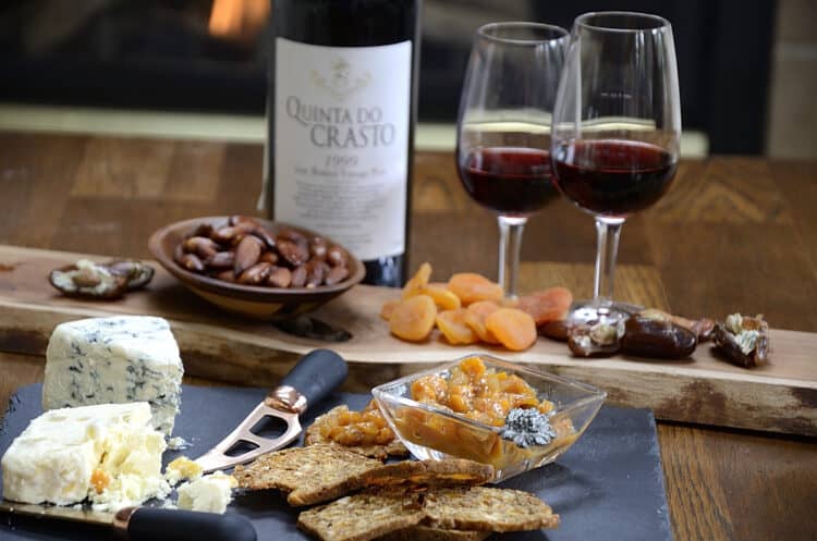 Bottle of Quinta Port with two glasses beside a charcuterie board with dates, apricots, blue cheese, chuntey and crackers.