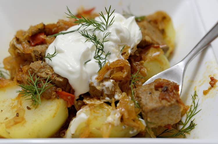 Close up of Transylvanian Goulash in a bowl over yellow potatoes, garnished with sour cream and fresh dill.