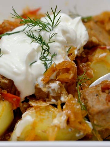 Close up of Transylvanian Goulash in a bowl over yellow potatoes, garnished with sour cream and fresh dill.