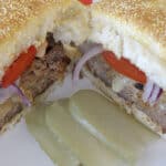 Juicy burgers filled with melted cheese, caramelized onion and bacon, on a bun on a summery plate with dill pickle slices.